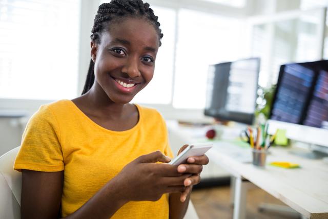 Young African American female graphic designer smiling while using a mobile phone in a modern office. Ideal for content related to technology, workplace diversity, professional women, and creative industries.