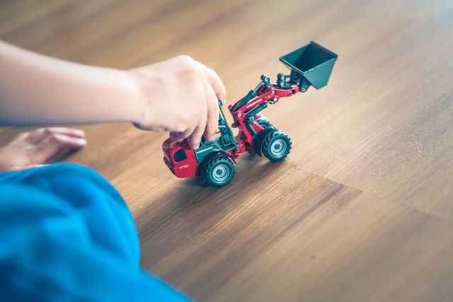 Close-up shot of a child's hand playing with a red toy tractor on a wooden floor. Suitable for concepts related to childhood, playtime, children's toys, and indoor activities. Ideal for use in parenting blogs, family lifestyle articles, and children's toy advertisements.