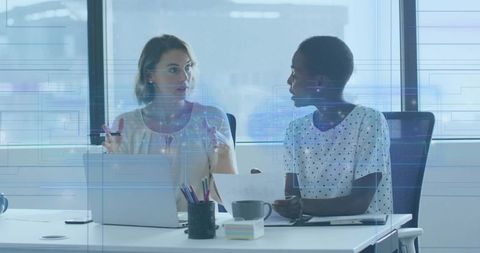 Female coworkers collaborating over laptop and documents at sunlit modern office desk