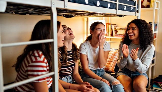 Laughing teenage girls chatting under bunk bed in cozy dorm room, casual friendship moment