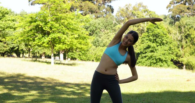 Woman engaging in a stretching exercise in a sunlit park with lush greenery and trees in the background. Perfect for use in health and wellness blogs, fitness guides, outdoor activity promotions, and summer exercise campaigns. Emphasizes an active and healthy lifestyle in a natural setting.