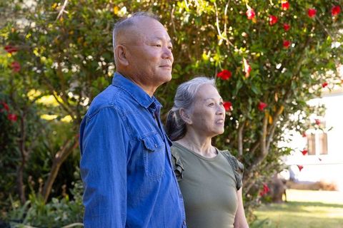 Serene senior couple standing in sunlit garden gazing at red blossoms and lush greenery