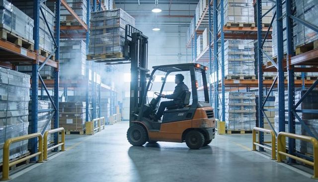 Forklift operator lifting pallet in high-bay warehouse aisle for logistics distribution