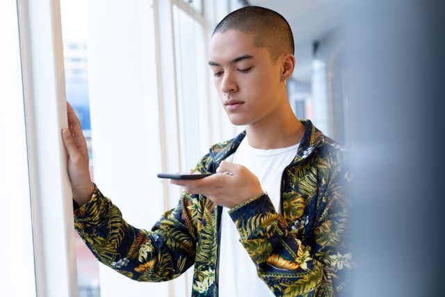 Young businessman standing near window using mobile phone in modern office. Ideal for corporate communications, technology usage, modern work environments, and business lifestyle themes.