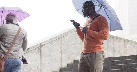 African american man holding umbrella and checking phone on rainy urban stairs