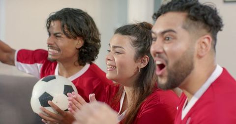 Friends cheering during soccer match at home wearing red jerseys on couch game day