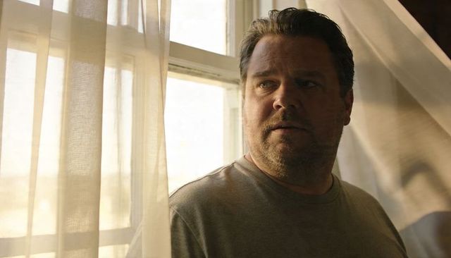 Middle-aged man gazing through window with golden-hour light and sheer curtains