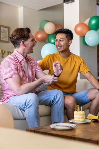 Young same-sex couple celebrating birthday at home, adjusting watch beside cake and balloons
