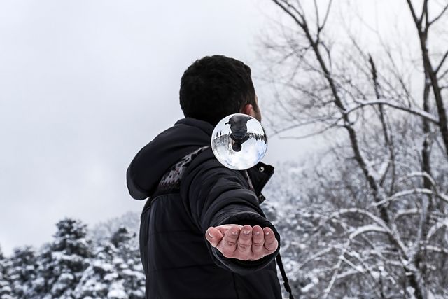 Man dressed in winter clothing holds a crystal ball in a snowy forest, with trees covered in snow. Reflection in the ball creates an artistic effect. Useful for concepts of magic, winter wonderland, serenity, and nature photography in wintry conditions.