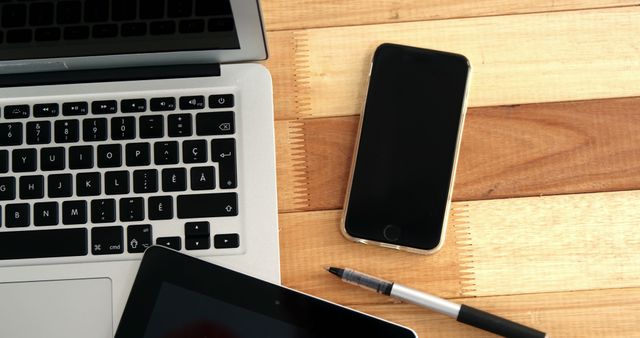 Scene depicts a modern workspace featuring a laptop, smartphone, tablet, and pen on a wooden desk. Ideal for themes related to digital work, remote productivity, technology use, and modern office environments.