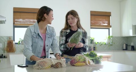 Two women unpacking groceries at bright modern kitchen island with reusable mesh bags