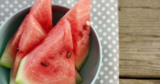 Close-up of fresh watermelon slices in a light blue bowl placed on a polka dot napkin with a wooden table background. Suitable for use in summer-themed blogs, healthy eating articles, and refreshing recipe ideas.