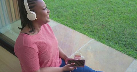 African american woman relaxing on doorstep wearing headphones holding smartphone and mug