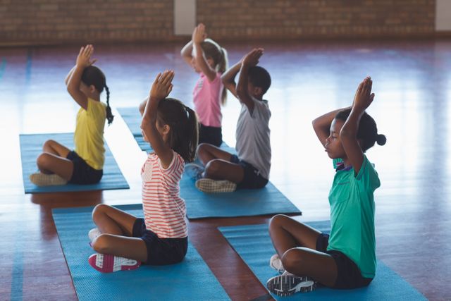 Children sitting on yoga mats meditating during a yoga class in a school gym. They are practicing mindfulness and relaxation techniques. This image can be used for educational materials, promoting children's fitness and wellness programs, or illustrating articles on the benefits of yoga and meditation for kids.