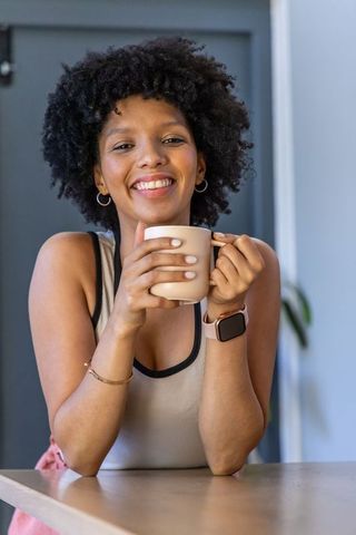 Smiling african american woman leaning on counter holding beige mug wearing smartwatch