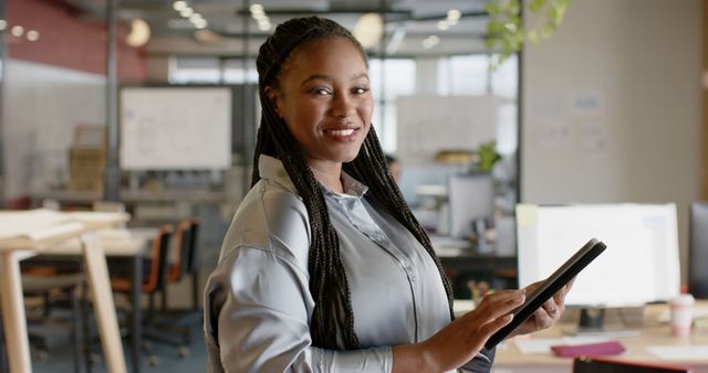 Businesswoman with braided hair smiling while using digital tablet in a modern office environment. Perfect for workplace diversity promotions, business magazines, corporate websites, and advertisements portraying modern technology in professional settings.