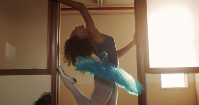 Side view of a biracial female ballet dancer practicing in an empty warehouse, standing in a distressed doorway, stretching.