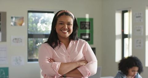 Smiling african american teacher crossing arms in bright classroom with student working