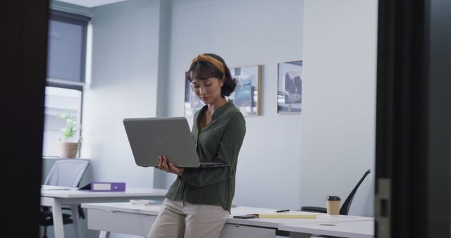 Young woman using laptop while standing in modern office. Ideal for use in business presentations, corporate training materials, professional development brochures, or workplace productivity blogs. Depicts casual work environment emphasizing modern technology and professional attire.