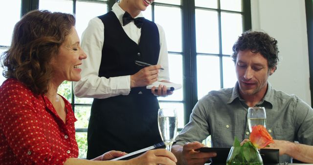 Couple at restaurant table interacting with smiling waiter taking order. Useful for concepts related to restaurant service, dining out, hospitality industry, and customer service. Can be used in marketing materials for restaurants, hospitality training guides, or lifestyle blogs discussing dining experiences.
