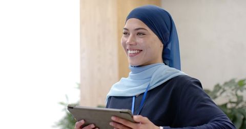Friendly muslim receptionist wearing blue hijab holding tablet, modern office smiling