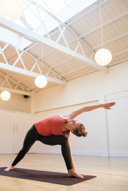 Woman practicing extended side angle pose on yoga mat in bright, spacious studio with natural light. Ideal for promoting yoga classes, fitness routines, healthy lifestyle, and wellness programs. Can be used in articles, blogs, and social media posts about yoga, exercise, and mental well-being.