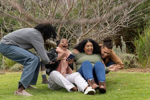 African american multigenerational friends laughing and holding smartphone while sitting on grass