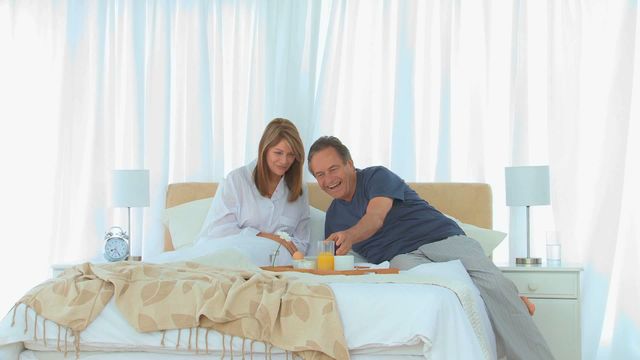 A couple is enjoying a relaxed morning as they dive into breakfast, adding a touch of happiness and intimacy to their day. This is ideal for showcasing retirement lifestyle, wellness, couples' time, and gentle morning routines in a cozy bedroom setting. Perfect for use in lifestyle blogs, relationship articles, and home setting inspirations.