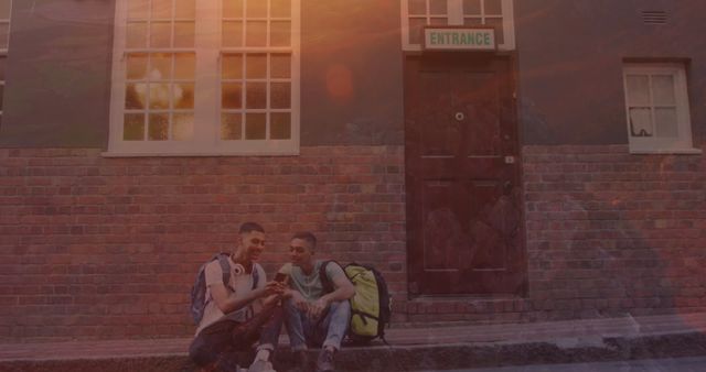 Two teenagers sit outside a brick building marked with an 'Entrance' sign. Both have backpacks and are using a smartphone. Suitable for themes of friendship, student life, outdoor activities, and casual daily moments.