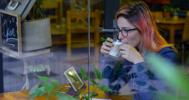 Young woman wearing glasses and stylish attire enjoying a hot drink in a cozy cafe with modern decor and plants, reflecting urban living. Ideal for lifestyle blogs, social media posts about cafe culture or modern relaxation, and advertisements for cozy coffee shops or eyewear brands.