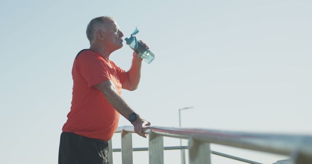 Senior man in red shirt drinking water from a bottle outdoors after a workout. He is seen resting by taking a break and staying hydrated, emphasizing the importance of healthy habits and outdoor activities. Perfect for use in health and wellness articles, fitness promotions, and advertisements focusing on staying active at any age.