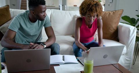 Couple working together on laptops at home sofa with notebook, papers and green smoothie