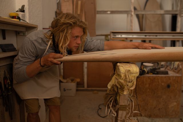 Caucasian male surfboard maker with long blonde hair wearing an apron, closely inspecting one of the surfboards being made in his workshop, with equipment in the background.