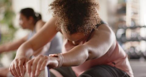 Female workout partners stretching and reaching foot on yoga mat wearing smartwatch in gym