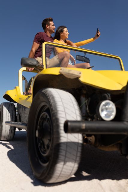 Happy caucasian couple sitting on beach buggy in the sun smiling and taking selfie. beach stop off on romantic summer holiday road trip.