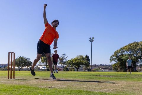 Fast bowler delivering ball midair on community cricket pitch under clear blue sky