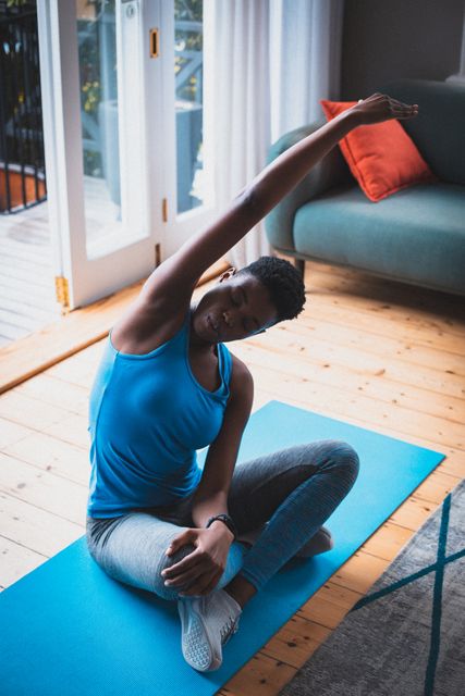 African American woman sitting on a yoga mat in a living room, stretching her arm overhead. Ideal for promoting home workouts, fitness routines, healthy lifestyle, and indoor exercise during quarantine or isolation. Useful for articles, blogs, and advertisements related to fitness, wellness, and home exercise.