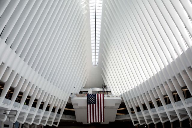 Modern architectural interior featuring impressive symmetrical structure with American flag hanging in center. Skylight adds natural light to white space, creating a futuristic and clean atmosphere. Ideal for articles or posters related to architecture, design inspirations, urban exploration, American landmarks, or patriotic themes.
