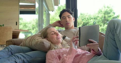 Relaxing diverse couple lounging on couch with tablet in modern sunlit living room