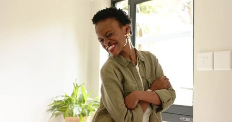 Smiling woman hugging herself near glass door with indoor plant and natural light