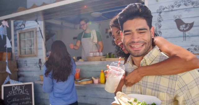 Friends enjoying food and drinks at an outdoor food truck. One friend giving a piggyback ride while holding a drink and food. Perfect for themes related to friendship, street food, outdoor dining, and casual gatherings.