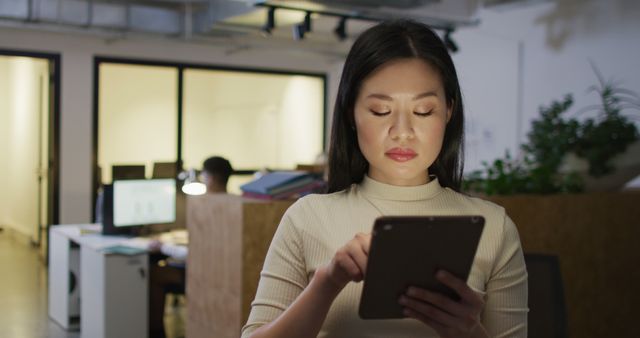 Asian woman using a tablet in a modern office environment. Evening setting, adding a sense of dedication and focus. Useful for themes related to technology, professional work, business environments, digital workflows, and evening work hours.