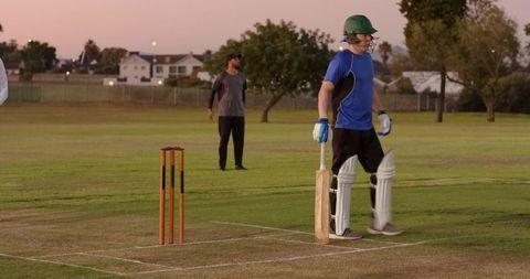Batsman standing at crease during dusk on suburban cricket pitch wearing green helmet