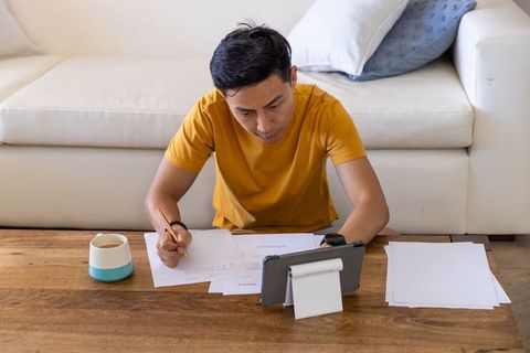 Remote worker writing bills and taking notes while referencing tablet at living room table