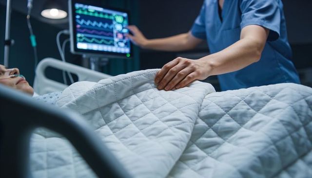 Nurse adjusting quilted blanket over hospital patient with cardiac monitor active