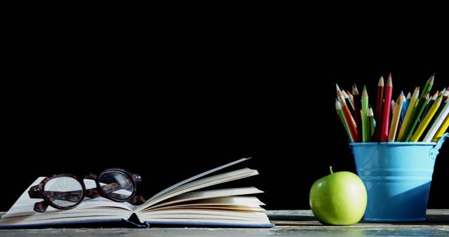 Open book with eyeglasses on top sitting on wooden desk next to a green apple and a blue bucket filled with colored pencils. Black background emphasizes items on the desk. Useful for educational themes, back to school promotions, study guides, and motivational content related to learning and reading.
