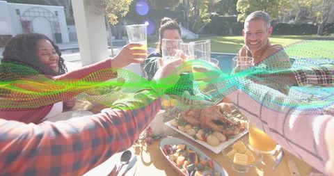 Friends toasting on sunlit patio around roast dinner by poolside, wearing casual sweaters
