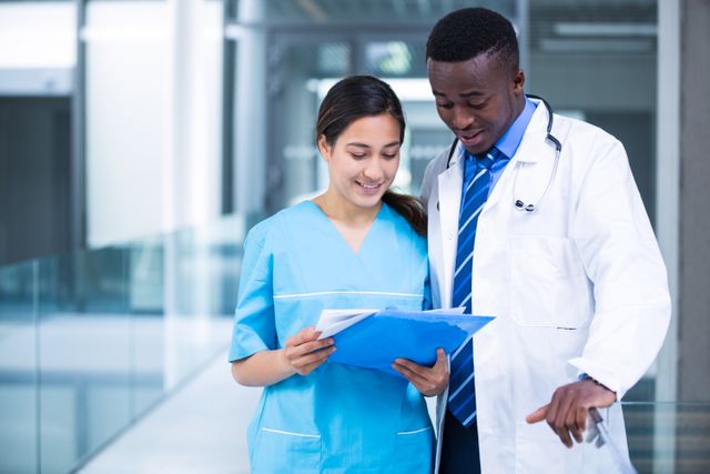 Doctor and nurse are reviewing a medical report in a hospital corridor. This image can be used to depict teamwork and collaboration in healthcare settings, medical consultations, and professional interactions among healthcare workers. Ideal for medical websites, healthcare blogs, and educational materials.