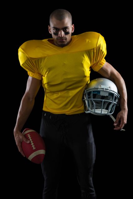 This image features a confident American football player holding a ball and helmet, dressed in a yellow jersey against a black background. Ideal for use in sports promotions, athletic advertisements, team posters, and articles about football or athleticism.