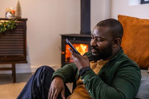 African american man using smartphone for voice message by cozy wood-burning stove